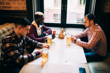 Technologies, online communication and internet addiction. Handsome Caucasian friends and their African American friend or partner enjoying fresh beer at pub and browsing social networks on mobiles