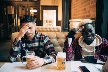 Handsome men's friends are sitting upset while drinking beer and cheering for sport team at the pub