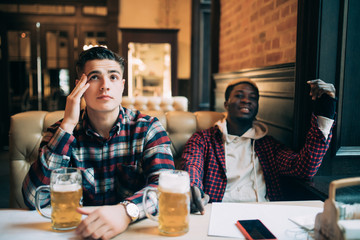 Upset male friends watching sport game or football match and drinking beer at bar or pub
