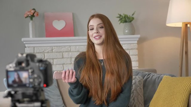 Young red-haired girl blogger, smiling talking on camera, using smartphone gestures with hands, home comfort in the background. 60 fps
