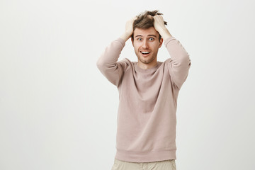 Indoor shot of funny handsome guy messing his hair while rubbing head, expressing devastation and acting crazy, standing over gray background. Guy is shocked after seeing home much work is left