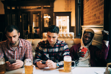 Technologies, online communication and internet addiction. Handsome friends and their African American friend enjoying fresh beer at pub and browsing social networks on mobiles