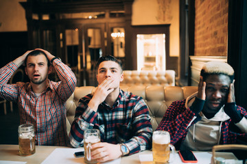 Handsome friends are sitting upset while drinking beer and cheering for sport team at the pub. Mens...