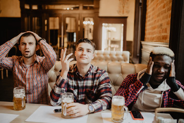 Handsome friends are sitting upset while drinking beer and cheering for sport team at the pub. Mens...