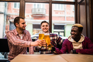 Cheers, my friends. Three multiracial handsome men are drinking beer, celebrating meeting and smiling.