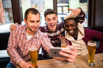 Three multiracial young men in casual clothes are taking selfie and drinking beer while sitting in pub