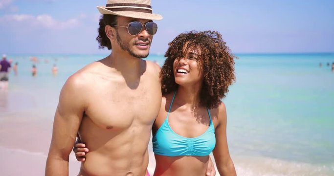 Happy young ethnic man and woman smiling and embracing on sandy shore at the ocean in sunny day.