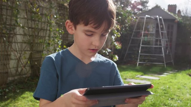 Young boy playing on his touchscreen tablet in the garden on a bright sunny day