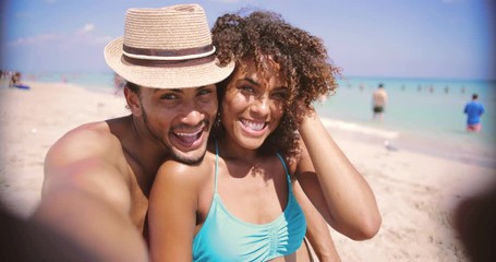 Laughing happy ethnic young man and woman looking at camera and taking selfie on sandy beach at the ocean. - Powered by Adobe