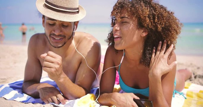 Happy young ethnic couple lying together and listening to music with earphones on beach at the ocean.