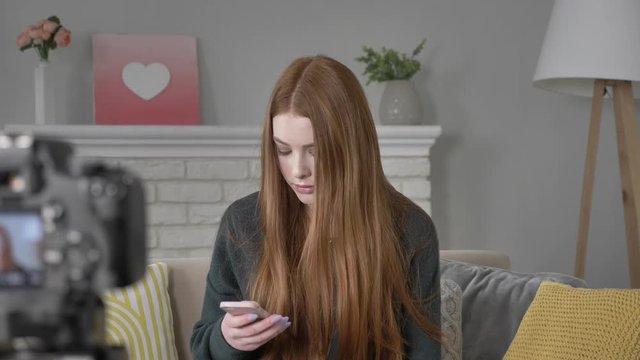 Young red-haired girl blogger at the camera, using a smartphone, texting, typing, home cosiness in the background. 60 fps