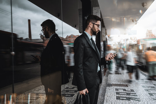 Handsome young caucasian bearded man employer in a formal suit is standing next to a bus stop and texting message on the smartphone, surrounded by city fussiness, walking people, and passing buses