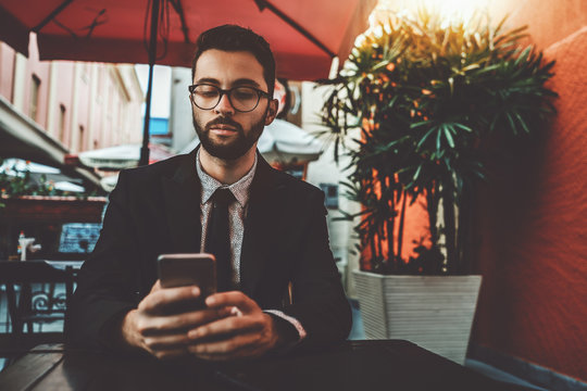 Serious businessman with the beard and formal suit, in the glasses, is sitting alone in an outdoor cafe next to his office and waiting for his lunch while using the smartphone to talk with colleagues
