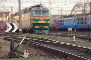 Fototapeta premium Old manual hand operated railroad switch with old locomotives and railway depot on background at sunset, selective focus/ Transportation, railroad traffic management concept/ Arrow switching/ 