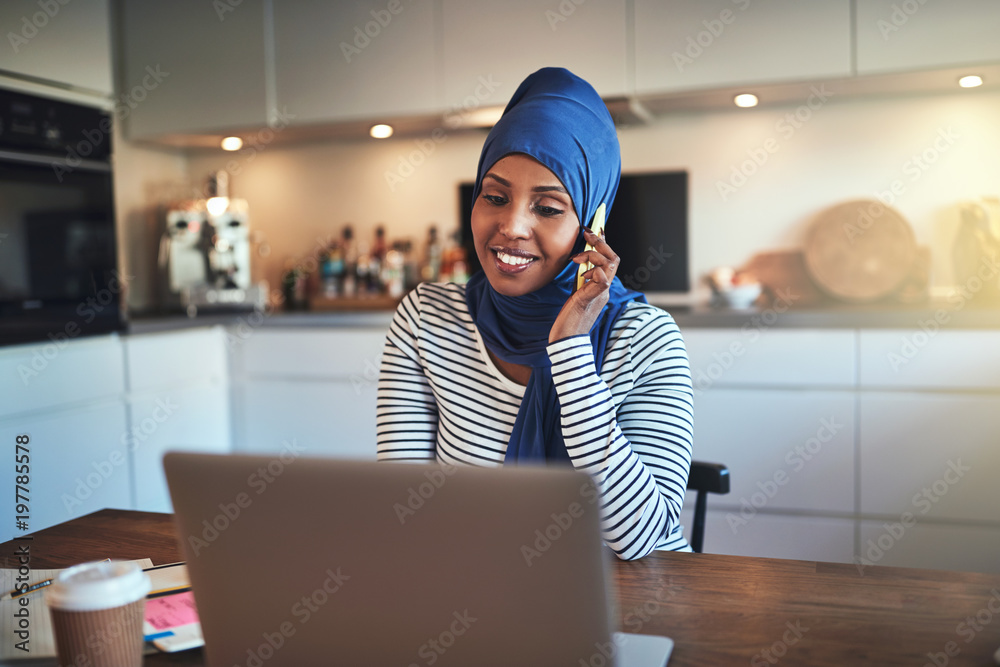 Wall mural Smiling Arabic entrepreneur talking on a cellphone in her kitche