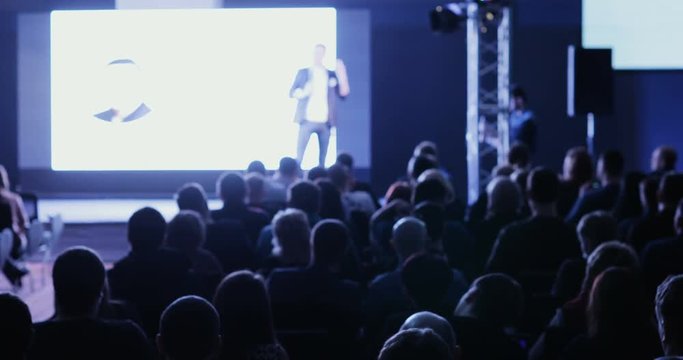 lecturer tells and shows presentation on projective screen to packed house of listeners