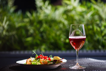 A plate with a beautiful appetizing salad of fresh vegetables, shrimps, quail eggs and lingonberries is on the table with a drink in a wine glass.