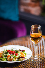 A plate with a beautiful appetizing salad of fresh vegetables, shrimps, quail eggs and lingonberries is on the table with a drink in a wine glass.