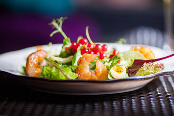 A plate with a beautiful appetizing salad of fresh vegetables, shrimps, quail eggs and lingonberries is on the table with a drink in a wine glass.