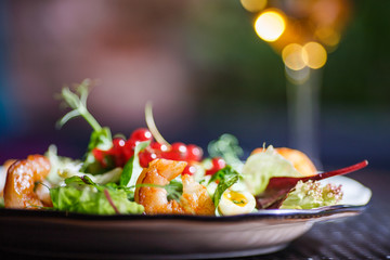 A plate with a beautiful appetizing salad of fresh vegetables, shrimps, quail eggs and lingonberries is on the table with a drink in a wine glass.