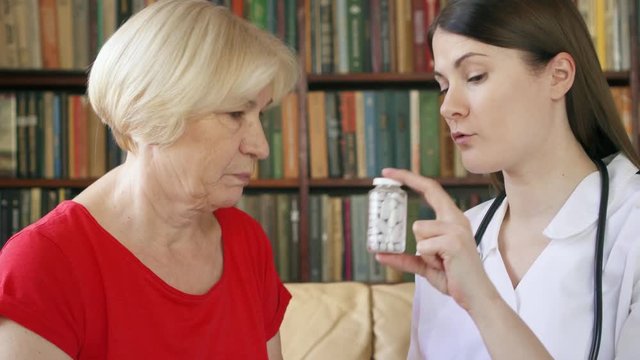 Female professional budding doctor in white coat with stethoscope at work. Young woman physician talking to sick senior female patient at home consulting about new drug. Showing bottle of pills