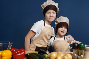 Food and nutrition concept. Isolated shot of two cheerful little boys siblings posing in kitchen while going to cook vegetarian lasagne for dinner together, wearing identical aprons and chef hats