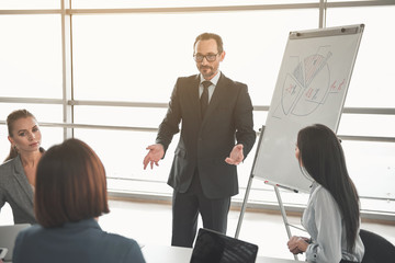 Young manager in suit reaching out to the audience in front of him while standing close to whiteboard with chart on it
