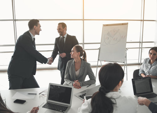 Team of executives sitting at meeting. Two men standing and shaking hands with happiness