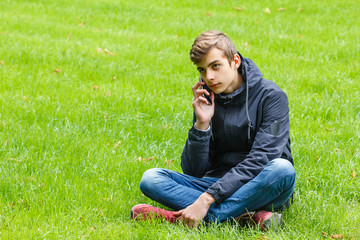 Male student talking on a phone seated on a grass in the city park