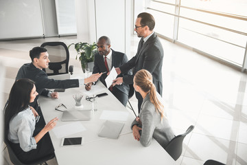 Group of people sitting during working meeting with chief. Boss standing with paper in hand while male participant pointing at it
