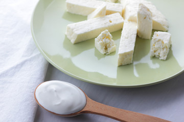 Cheese and sour cream, feta in a green plate, soft cheese on a white background, wooden spoon with sour cream, French breakfast, blue napkin, dairy products on a light background, copy space