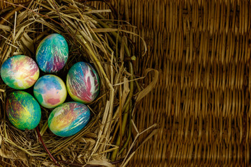 Many multi-colored Easter eggs are in the nest of grass and branches. Nest lies on the table background made of braided willow branches.