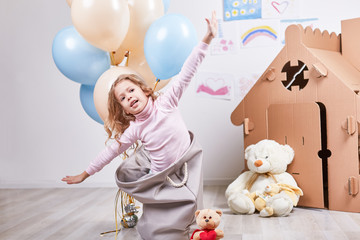 Fototapeta na wymiar Portrait of happy kid imagines she is airplane pilot. Child has fun in nursery room with toy house and teddy bear