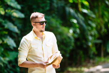 Young man in sunglasses casual clothes holding ylellow hat in hands having fun in sunny day. Tropical background.