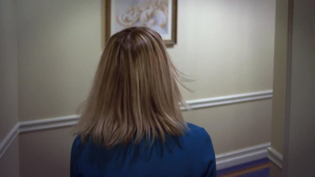 Young blonde woman in blue dress walking on hallway corridor at guest hotel. Close up woman walking in room number in corridor back view