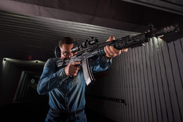 An adult man in jeans clothes, headphones and glasses, holding a Kalashnikov rifle with an optical...