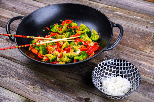 wok with vegetables, bowl of rice and chopsticks on wooden ground