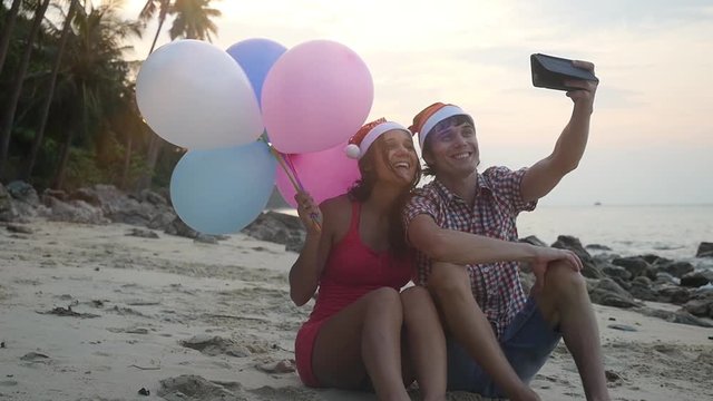 Happy funny couple sit on the beach with multicolored balloons on Christmas travel holidays taking selfie picture with smartphone wearing santa hat during sunset in slow motion. 1920x1080