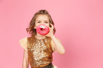 Surprised little girl in glittering dress standing with donuts posing on pink background.