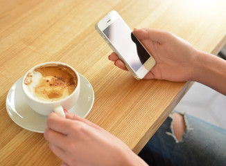 A mock-up of hands with a white mobile phone with an empty black screen. A girl is using a smartphone while sitting in a cafe. Conceptual photography. A cup of coffee. Lunch break. Communication. 