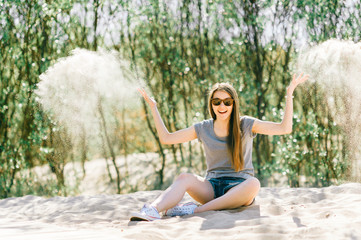 Fototapeta na wymiar Amazing young girl siiting on beach and looking at camera. Fashion portrait of beautiful female playing with sand beyond sea. Pretty cheerful person smiling face. Traveler relaxing at nature outdoor.