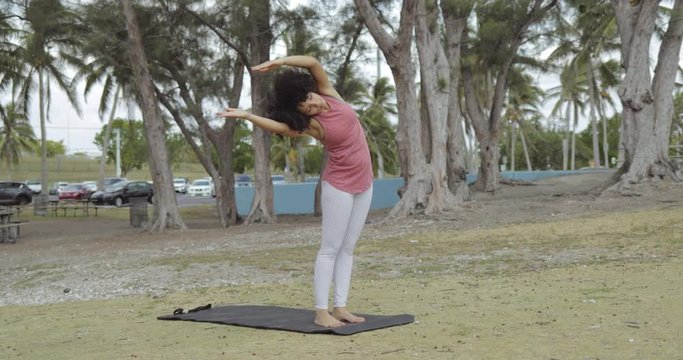 Young sportive African-American woman standing on mat in park and bending on sides practicing yoga. 