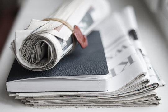 Business concept - folded newspapers stacked in a pile, notebook with leather cover and rolled newspaper with seal. Daily papers with fresh financial news and book for notes on the desk, close up