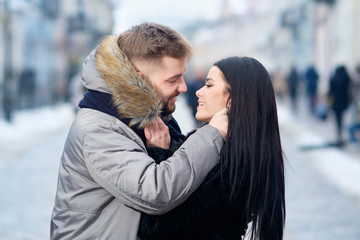 Fototapeta premium young happy lovely caucasian couple hug and kiss each other while walking in the street of town in winter under snowfall