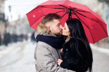 young teen couple hug each other standing in center of street uner red umbrella in winter under snowfall, looking in eyes to each other and smiling