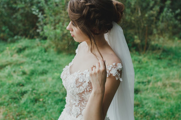Naklejka premium Beautiful bride with elegant simple hairdo is standing in the forest. Low open back white dress with flowers and tulle veil. Stylish hairstyle in outdoors romantic wedding photo. Rustic details.