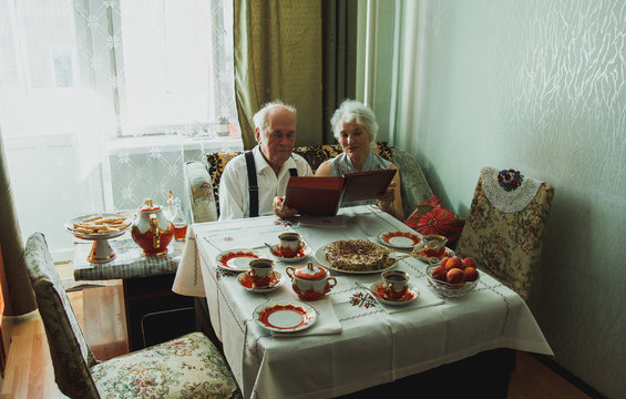 Old couple is flipping the photo album at home. Grandmother and grandfather at golden wedding anniversary celebration in vintage dining room near the table with cups, cake and tea. Happy grandparents.