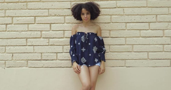 Content coquettish girl with short curls posing in trendy shirt and shorts standing with hands on waist looking at camera. 