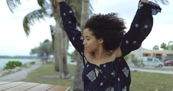 Beautiful black girl in stylish blouse holding hands behind head and enjoying fresh wind with eyes closed sitting on beach. 