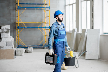 Builder in working uniform with protective helmet standing with instruments at the construction site indoors